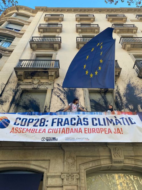 protestors and scientists stand on the black paint splattered balcony of the european union offices with a banner that reads  cop28 fracàs climático assemblea ciutadana europea ja